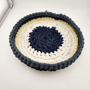 May include: A round crocheted basket with a white, yellow, and navy blue design. The basket is made of thick yarn and has a textured surface.