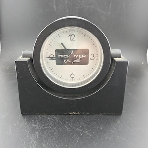 May include: A black analog clock with a white face and black hands. The clock face has the number 12 at the top and the number 3 on the right. The clock is set in a black rectangular base. The text "DICK YER original" is visible on the clock face.