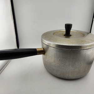 May include: A vintage aluminum saucepan with a black handle and a lid. The saucepan has a brass accent on the handle.