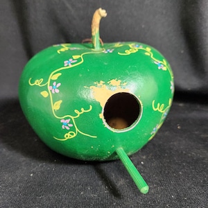 May include: A green gourd birdhouse painted with a vine and flower design. The birdhouse has a round opening and a green stick for a perch.