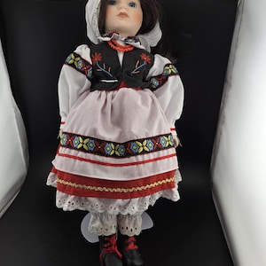 May include: A porcelain doll dressed in traditional Polish folk attire. The doll wears a white head covering, a black vest with floral embroidery, a pink skirt with red and gold trim, and black boots with red laces. The doll has blue eyes and brown hair.