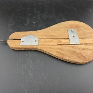 May include: A wooden tool with a teardrop shape, featuring a metal blade and two metal brackets. The wood has a light brown color, and the metal components are silver. The tool appears to be a specialized cutting or shaping instrument.