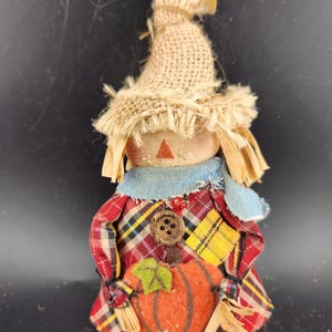 May include: A small, handmade scarecrow doll with a burlap hat, plaid shirt, and blue jeans. The scarecrow is holding a felt pumpkin.