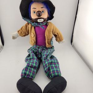 May include: A plush clown doll with a black hat, blue hair, a brown beard, and a plaid outfit. The doll is sitting with its legs crossed.