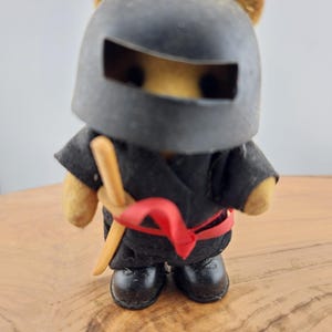 May include: A plush brown bear dressed as a ninja, wearing a black helmet with a visor, a black robe, a red sash, and holding a wooden sword.