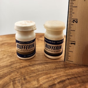 May include: Two small, vintage Bufferin analgesic tablet bottles. The white bottles have a blue and gold label with the brand name and text. A wooden ruler is next to the bottles.