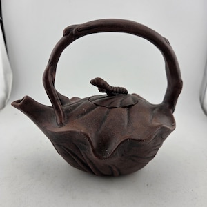 May include: A brown ceramic teapot with a leaf-like design and a curved handle. The teapot has a spout and a small decorative element on the lid. The handle is arched and appears to be made of the same material as the body.