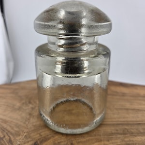 May include: A clear glass jar with a silver-colored lid. The jar has a rounded body and a slightly textured surface. The lid is dome-shaped. The jar is sitting on a wooden surface.