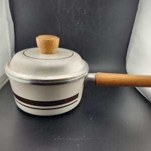 May include: Cream-colored saucepan with a lid and wooden handle. The pan has a brown stripe around the middle and a wooden knob on the lid. The handle is made of wood and metal. A metal loop is attached to the end of the handle.
