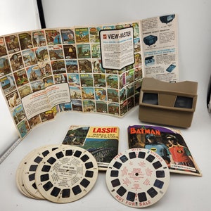 May include: A vintage View-Master projector with a brown and beige color scheme. The projector is surrounded by several View-Master reels, including "Star Trek", "Lassie Rides the Log Flume", and "Batman". The reels are colorful and feature images of the characters from the respective shows. The text "FREE WITH VIEWER" is printed on the Batman reel.