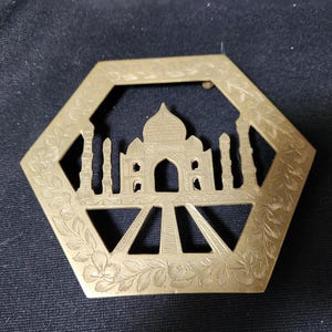 May include: A gold-colored metal coaster with an intricate design of the Taj Mahal. The coaster is shaped like a hexagon and has a floral pattern around the edge.
