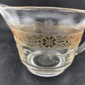 May include: Clear glass creamer with a handle and spout. The creamer features a decorative band with a floral design and etched leaf patterns. The glass has a slightly yellow tint.