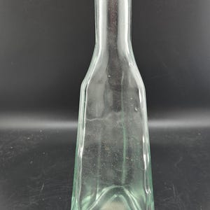May include: A clear glass bottle with a tall, slender neck and a square-shaped base. The body of the bottle features slightly concave sides and a smooth, transparent finish, tapering upward from the wide base to a narrow, circular opening at the top.