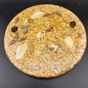 May include: A round, clear resin coaster with a variety of seashells, sand, and pearls embedded inside. The coaster has a light brown color and is perfect for adding a touch of the beach to your home decor.