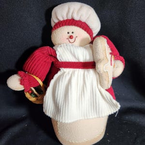 May include: A plush gingerbread woman doll wearing a red sweater, white apron, and a white baker's hat. She is holding a gingerbread cookie in one hand and a basket with greenery in the other.