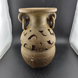 May include: A brown ceramic vase with a wide top and base, featuring intricate cut-out designs and two loop handles. The vase has a detailed, carved pattern around the base and a textured surface.