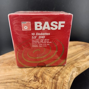 May include: A red box of BASF 3.5" 2HD diskettes. The box has the BASF logo and text in multiple languages, including "Two sided - High density" and "Certified 100% error free". The box is sitting on a wooden surface.