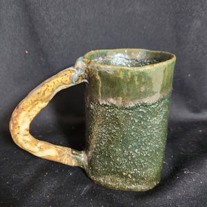 May include: A green and brown ceramic mug with a unique, textured finish and a handle that is a lighter brown color.