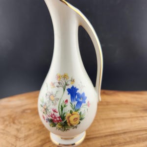 May include: A small white porcelain vase with a gold rim and floral design. The vase has a curved handle and a delicate floral pattern in shades of blue, yellow, pink, and green.