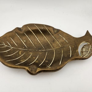 May include: A bronze-colored leaf-shaped ashtray with etched veins and a small leaf cutout. The ashtray has a shallow dish for holding items and is made of a metallic material. The design is detailed and decorative.