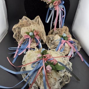 May include: A collection of vintage heart-shaped ring pillows and a heart-shaped display stand. The pillows are cream-colored, adorned with lace, pink and blue ribbons, and small floral arrangements. The stand is black and has a lace trim.