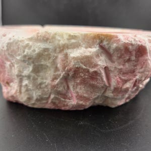 May include: A roughly hewn, circular stone bowl with a pink and white marbled appearance. The bowl's exterior has a textured, natural look, suggesting it is made from a type of stone or mineral. The color palette is soft and earthy.