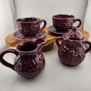 May include: A purple ceramic tea set with a grape design. The set includes a teapot, creamer, sugar bowl, and two teacups and saucers.