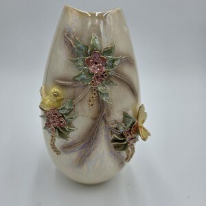 May include: Cream-colored decorative vase with a pearlescent finish. The vase is adorned with floral and insect embellishments in shades of pink, green, and gold. The vase has a unique, elongated shape.
