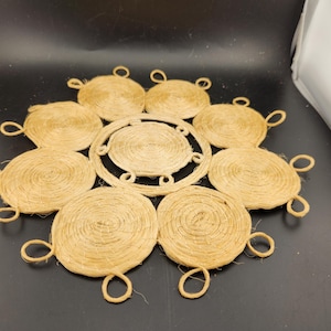 May include: A round, tan-colored, woven jute coaster with a circular design in the center. The coaster is made of multiple layers of jute rope, creating a textured and rustic look.