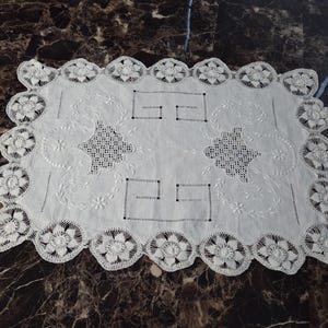 May include: A rectangular white linen doily featuring intricate cutwork embroidery, geometric Greek key patterns, and a scalloped border composed of crocheted floral motifs. The central fabric displays delicate needlework and openwork details.