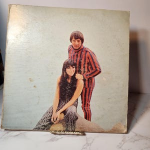 May include: Vintage album cover featuring a man and woman. The man wears a red and blue striped outfit. The woman is in a patterned dress. The album cover has a light blue background and is slightly worn.