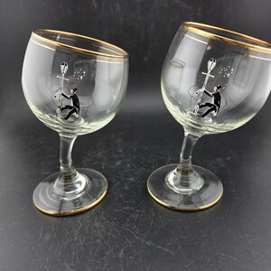 May include: Two clear wine glasses with gold rims and bases. Each glass features a black and white illustration of a person climbing a pole with stars. The glasses are on a dark surface.
