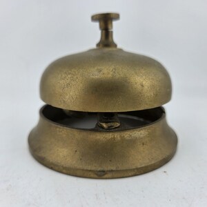 May include: A brass desk bell with a rounded dome top and a small cylindrical plunger button at the center. The metal surface has a weathered, aged patina with visible signs of wear and oxidation, featuring a wide, flared circular base.