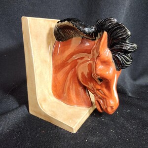 May include: A ceramic horse head bookend with a brown and black mane. The bookend is mounted on a beige base.