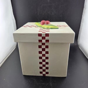 May include: A cream-colored, square ceramic box with a lid, decorated with a red and white checkered pattern. The top features a green leaf and red berry design. The box is a decorative item.