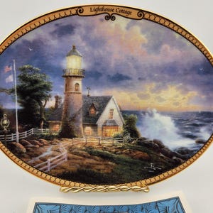 May include: Oval decorative plate featuring a coastal scene with a lighthouse and cottage. The plate has a gold rim and a gold stand. The lighthouse is lit, and the ocean waves are crashing against the shore. The text "Lighthouse Cottage" is at the top.