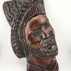 May include: A carved wooden bust depicting a man with a beard and hat. The sculpture is made of dark brown wood with visible grain and texture. The man's face has detailed features, including eyes, a nose, and a mustache. The bust is a decorative item.