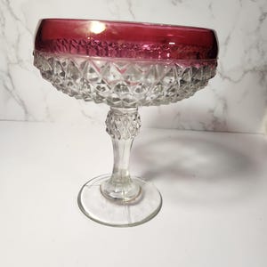 May include: A vintage glass pedestal bowl with a ruby red rim and diamond-cut detailing. The bowl sits on a clear, ornate stem and a circular base. This elegant piece is suitable for serving desserts or displaying decorative items.
