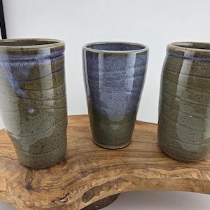 May include: Three handmade ceramic tumblers in shades of green, blue, and gray. The tumblers have a speckled texture and a glossy finish. They are displayed on a wooden surface, showcasing their unique shapes and colors.
