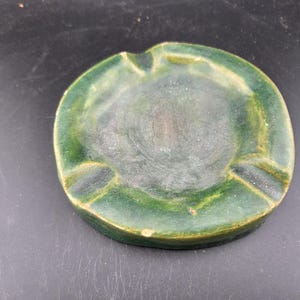 May include: A round, green ceramic ashtray with three notched rests for cigarettes. The ashtray has a circular design with a slightly raised edge and a dark green center.