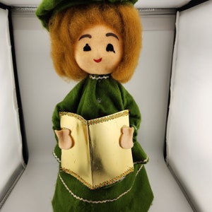May include: A green felt Christmas carol singer doll with a gold book and a green hat. The doll has brown hair and a smiling face.