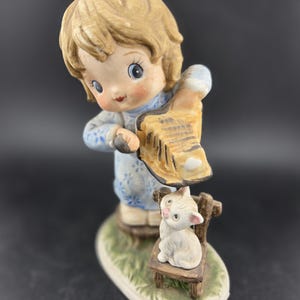 May include: A ceramic figurine of a young child in a blue dress, holding a musical instrument. A white cat sits on a small wooden chair. The figurine has a detailed, hand-painted design with a glossy finish.