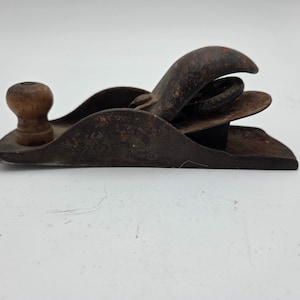May include: A small, antique hand plane with a wooden knob and a curved handle. The metal body shows signs of age and wear, with a rusty brown patina. This woodworking tool is designed for smoothing and shaping wood surfaces.