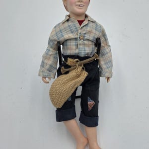 May include: A vintage doll of a young boy with red hair, wearing a plaid shirt, dark blue pants with patches, and a burlap sack tied around his waist. The doll has a painted face and is seated.