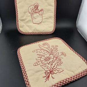 May include: Two square, cream-colored pot holders with red embroidered designs. One features a woman holding a baby, the other a floral bouquet. Both have a red and white checkered border.