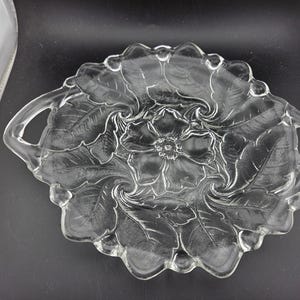 May include: Clear glass serving dish with a floral design. The dish has a scalloped edge and two handles. The floral pattern is in the center of the dish and is surrounded by leaves. The dish is approximately 10 inches in diameter.