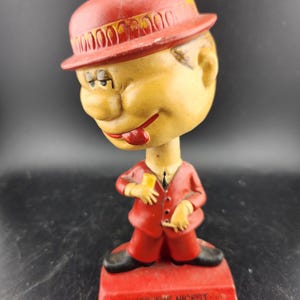 May include: A red bobblehead figurine of a man wearing a red hat and suit. The figurine is holding a cigarette in one hand and a drink in the other. The base of the figurine reads "You're the nicest of all my bad habits."