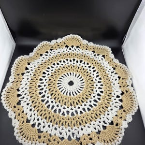 May include: A handmade, circular crochet doily in a layered design. The doily features alternating rings of beige and white yarn, creating a textured, openwork pattern. The outer edge has a scalloped trim.