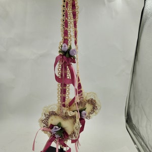 May include: Two white fabric hearts with lace trim and purple flowers hanging from a burgundy ribbon. The hearts are attached to a black stand.