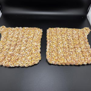 May include: Two handmade, square crochet potholders. The potholders are made with a thick, textured yarn in shades of yellow, brown, and cream. Each potholder has a small loop at one corner for hanging. The potholders are displayed on a black surface.